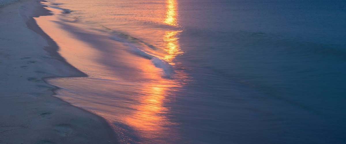 Ein Strand bei Sonnenuntergang, das ruhige Meer spiegelt die Farben des Himmels.