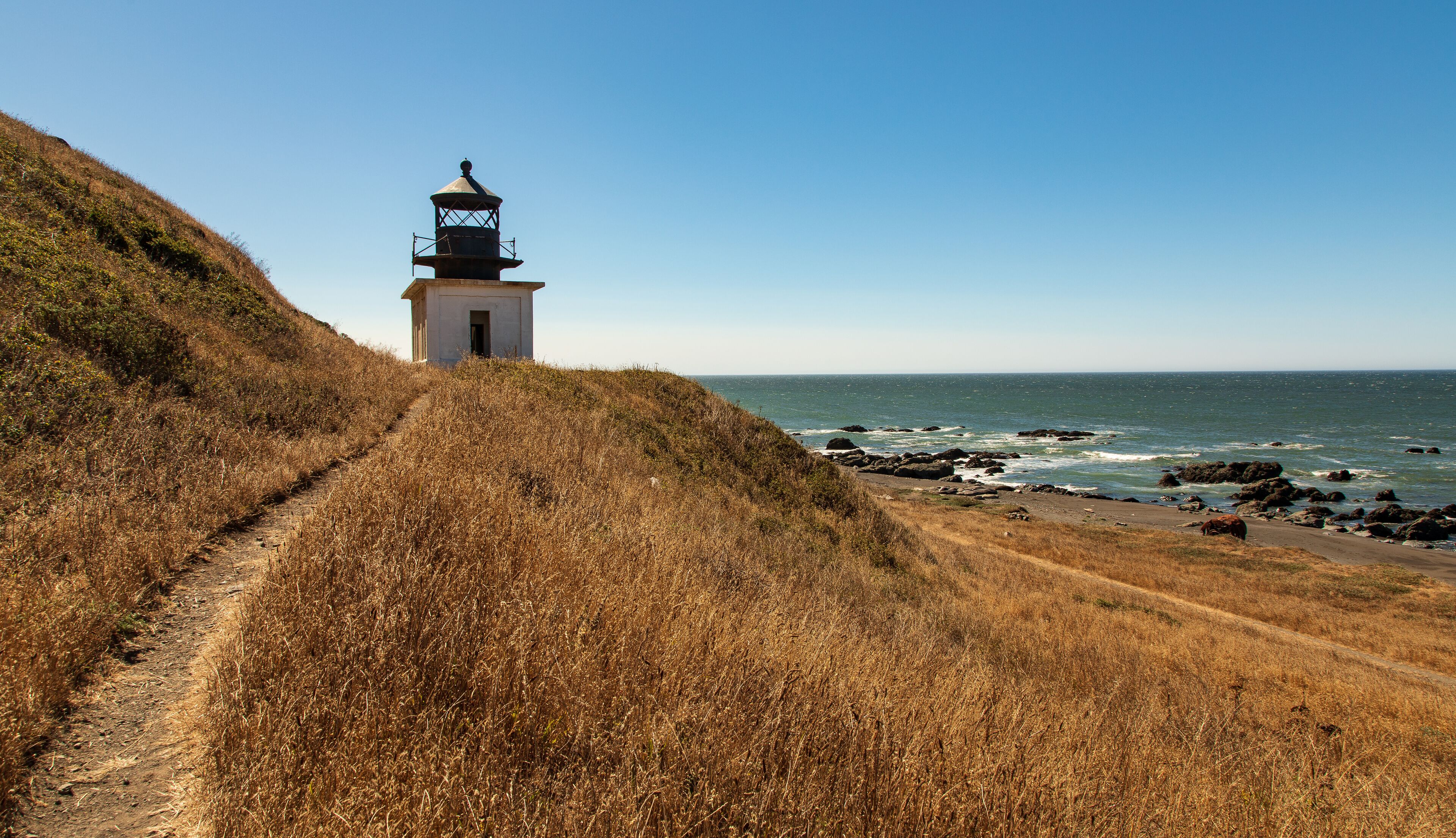 A coastal path leading to a lighthouse, surrounded by dry grass and rocky shoreline.