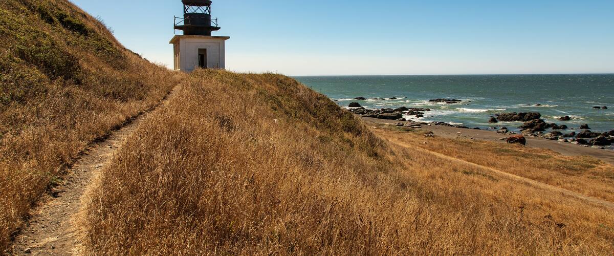 A coastal path leading to a lighthouse, surrounded by dry grass and rocky shoreline.