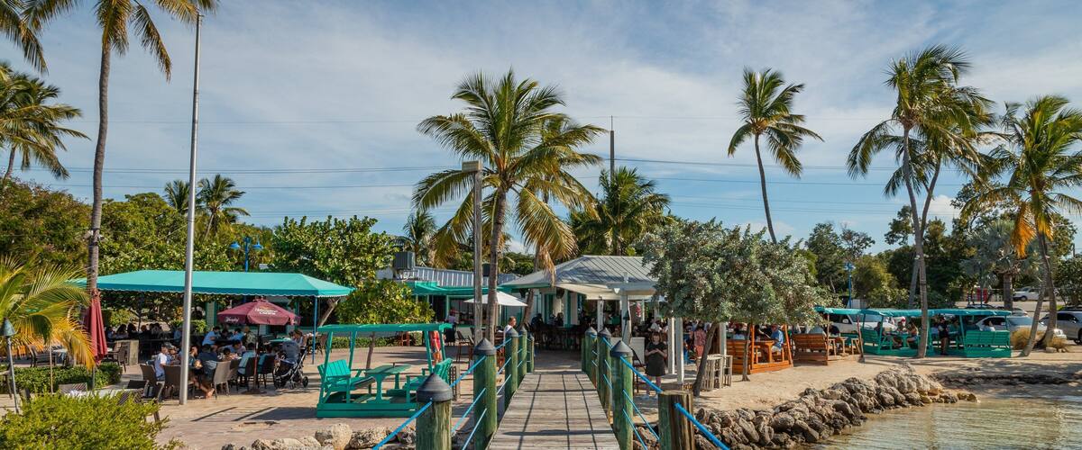 A wooden bridge over water, surrounded by palm trees and outdoor seating areas.