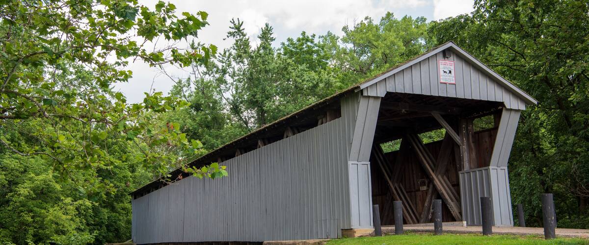 A covered bridge with a sign, surrounded by lush greenery and a small stream.