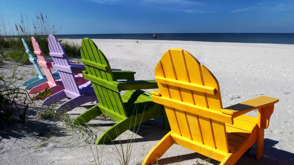Colorful Adirondack chairs on a sandy beach with the ocean in the background.