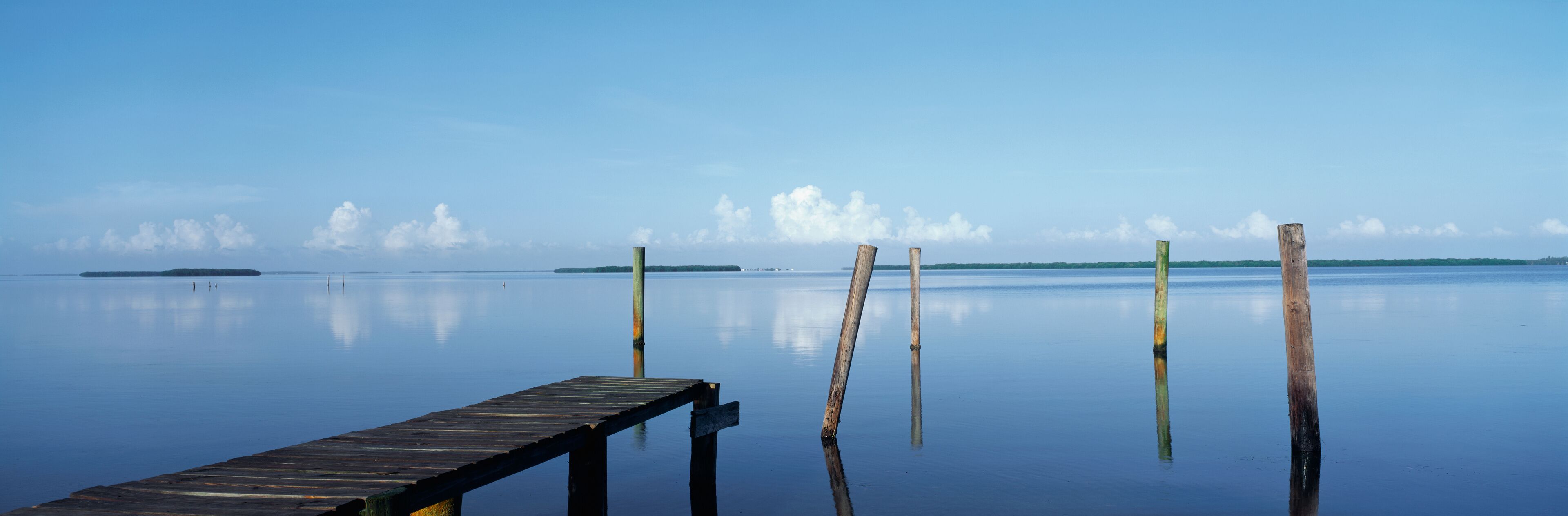 This is the morning view of Pine Island Sound. Its pier juts out from the left side with wooden pylons standing up out of the water near the shore.