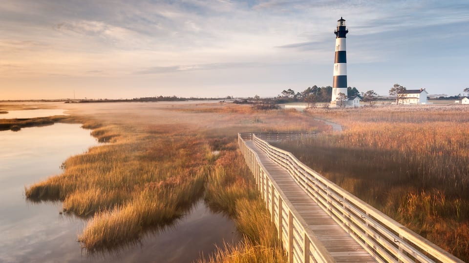 A lighthouse, a wooden boardwalk, and marshy landscape.