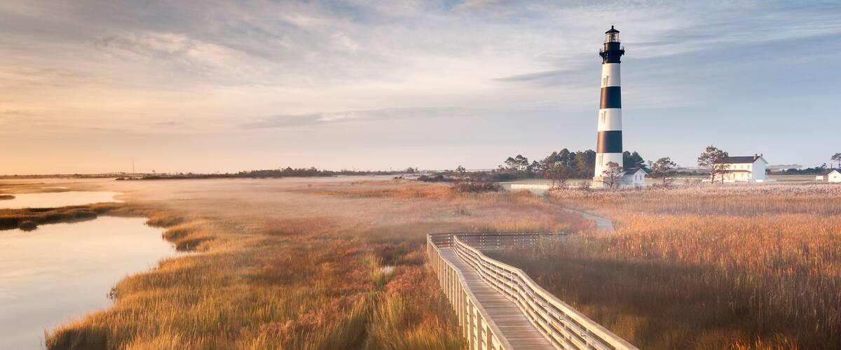 A lighthouse, a wooden boardwalk, and marshy landscape.