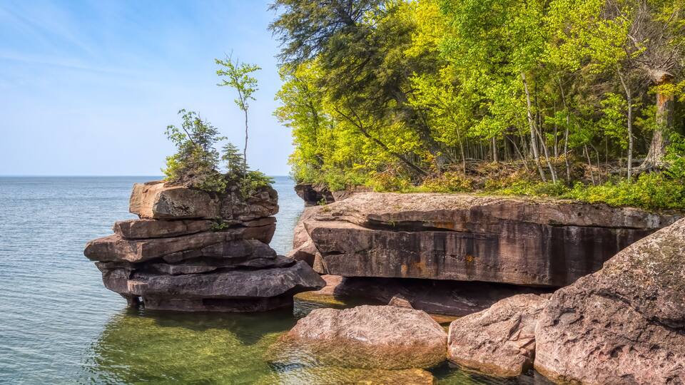 A rocky shoreline with clear water, surrounded by green trees and a clear sky.