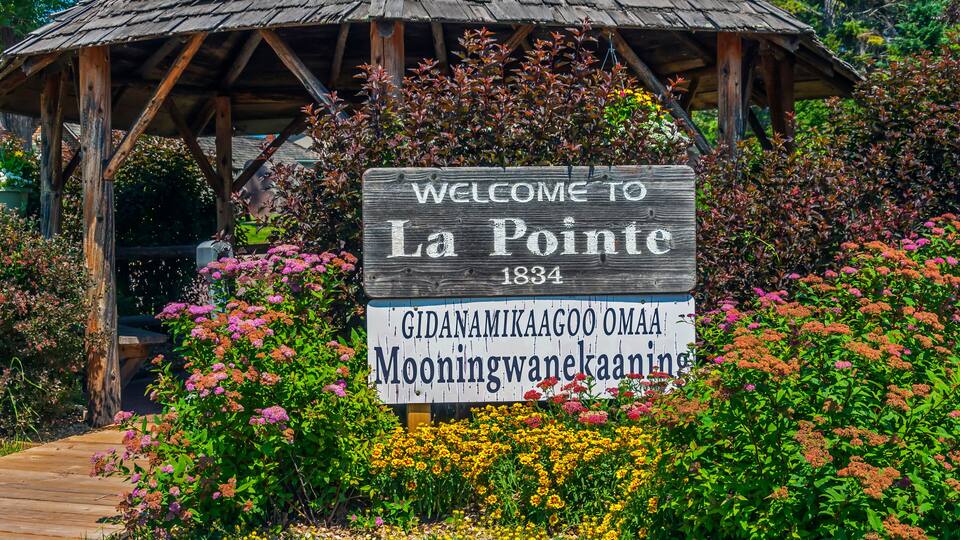 A wooden sign with 'WELCOME TO La Pointe 1834' and 'Gidanamikaagoo omaa Mooningwanekaaning' written on it, surrounded by colorful flowers and a wooden structure.