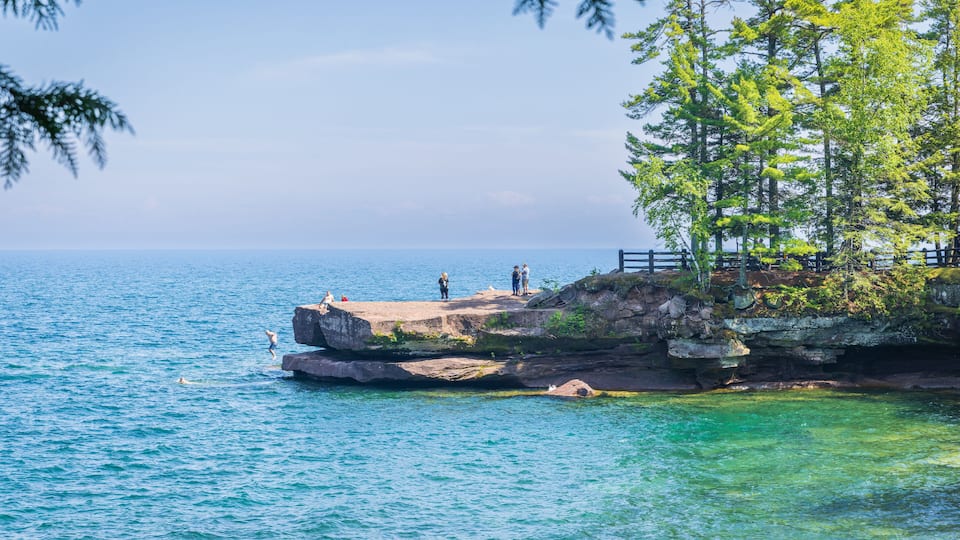 A rocky shoreline with clear blue water, a wooden pier, and lush green trees.