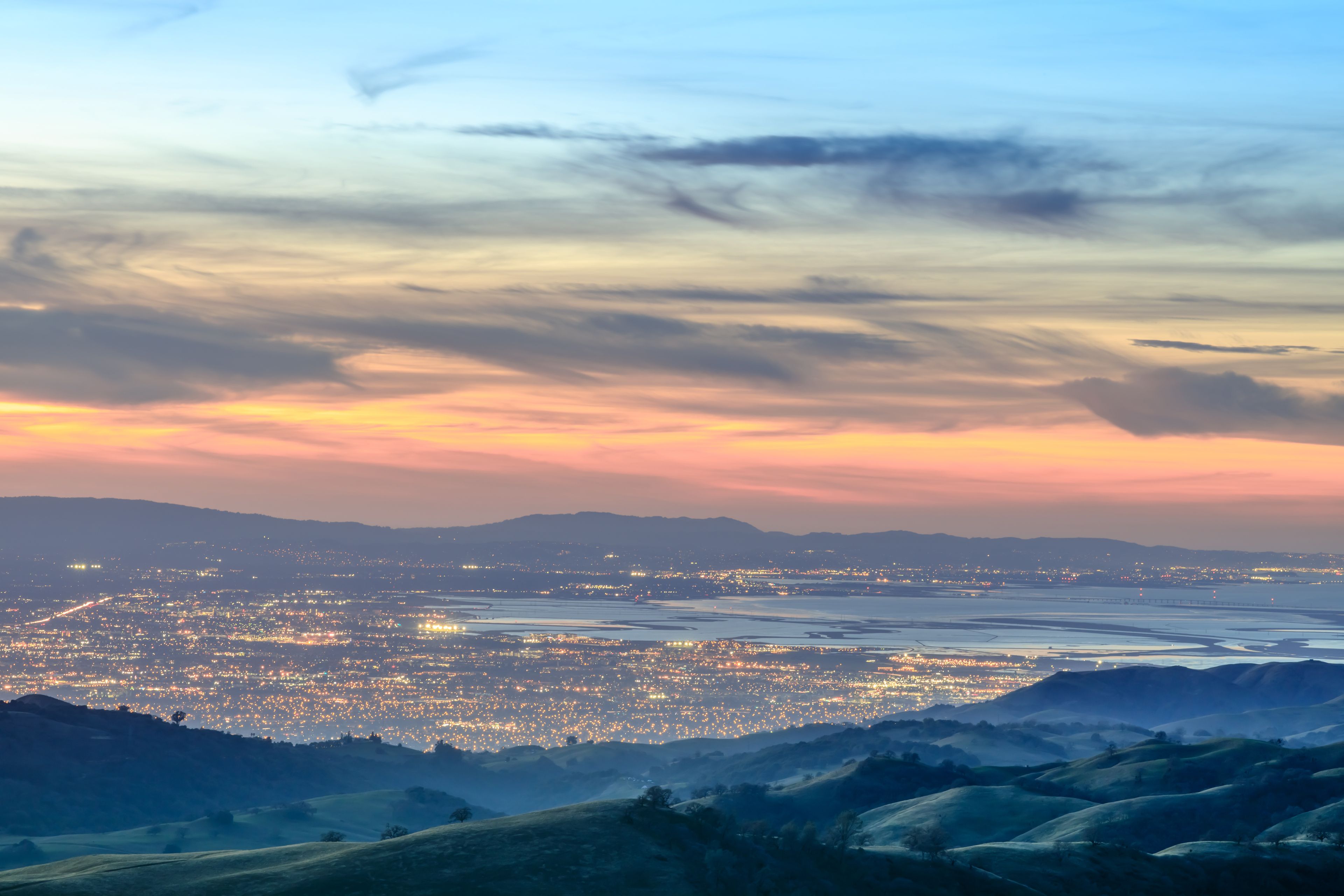 A cityscape at dusk with a sunset sky, illuminated city lights, and rolling hills in the foreground.