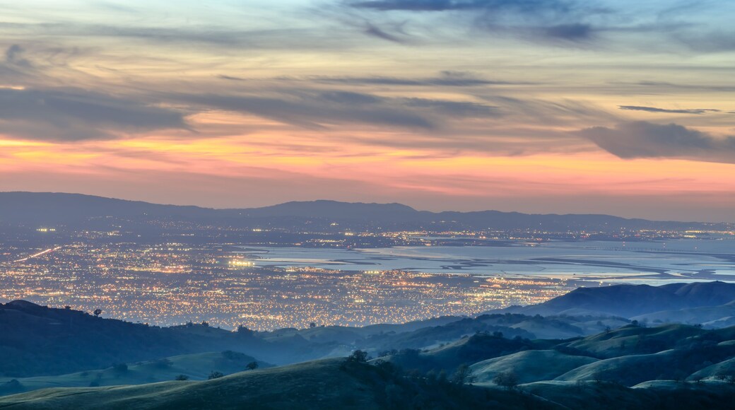 A cityscape at dusk with a sunset sky, illuminated city lights, and rolling hills in the foreground.