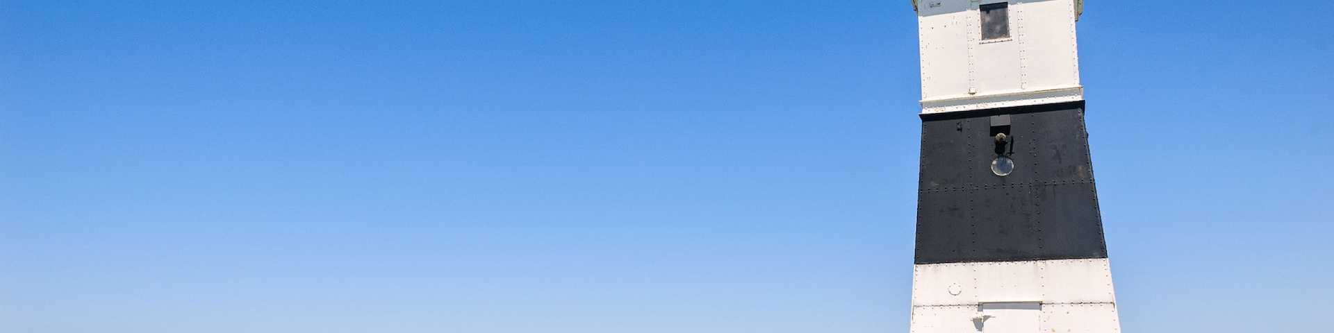 A tall, narrow lighthouse with a black top and white body, situated on a concrete pier extending into calm waters.