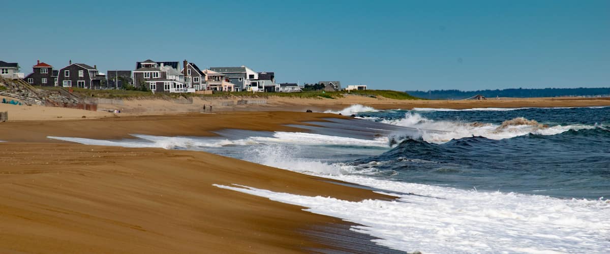 A sandy beach with waves crashing onto the shore. In the background, there are houses along the coastline.