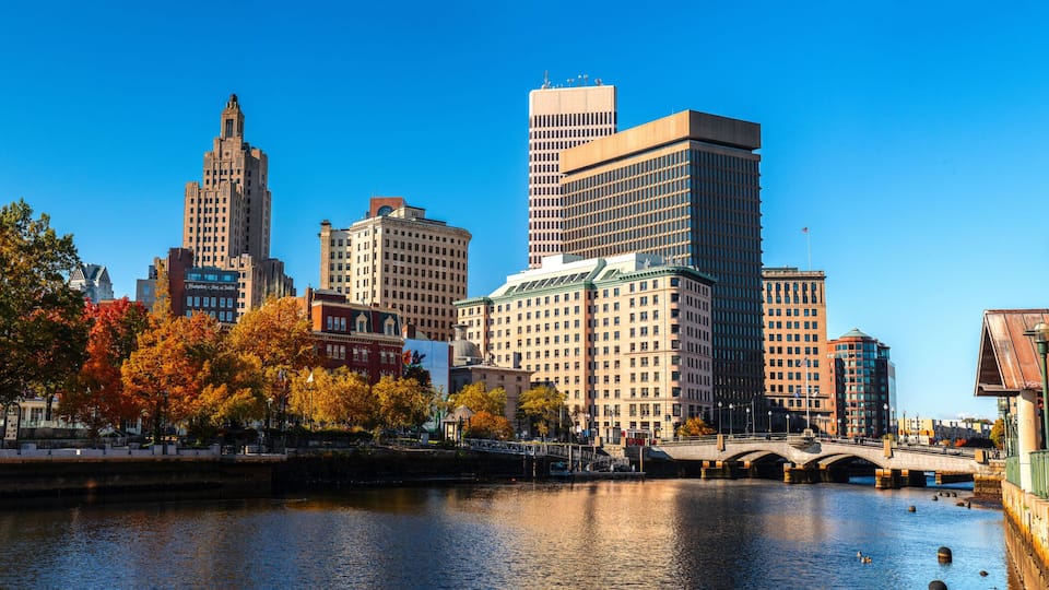 A river with a bridge, autumn trees, and a city skyline with various buildings.