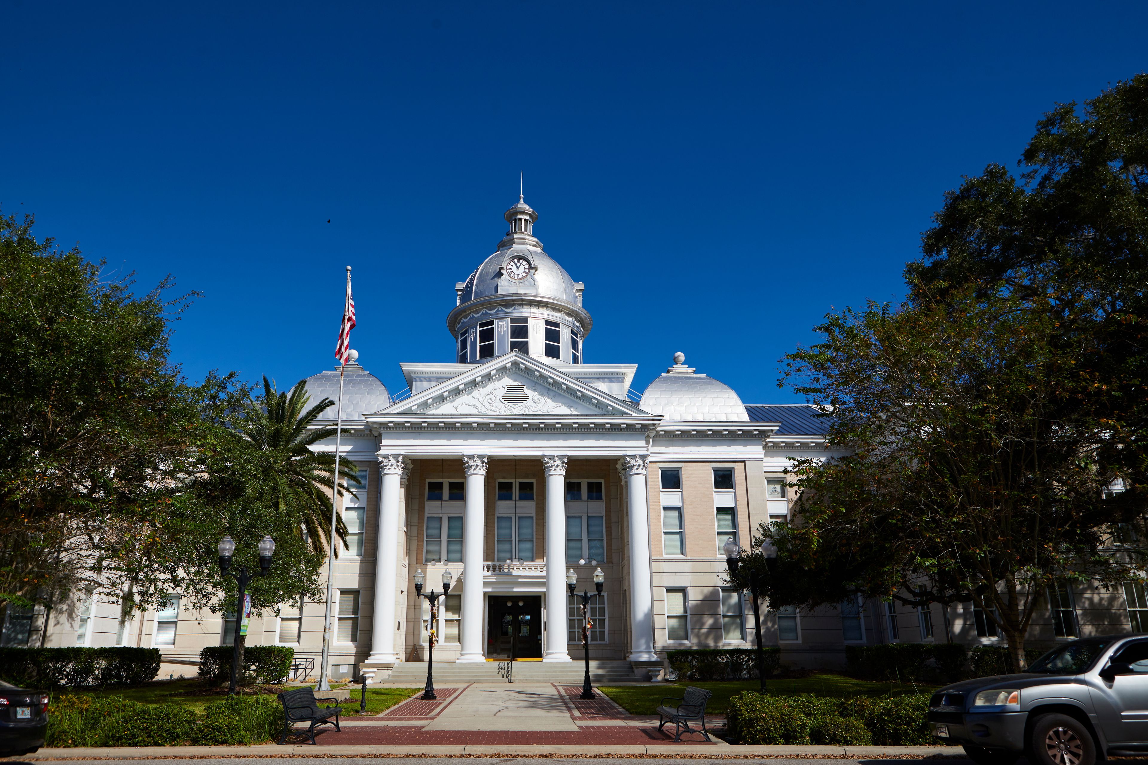 A large, classical-style building with a prominent dome and columns, surrounded by trees and a clear blue sky.