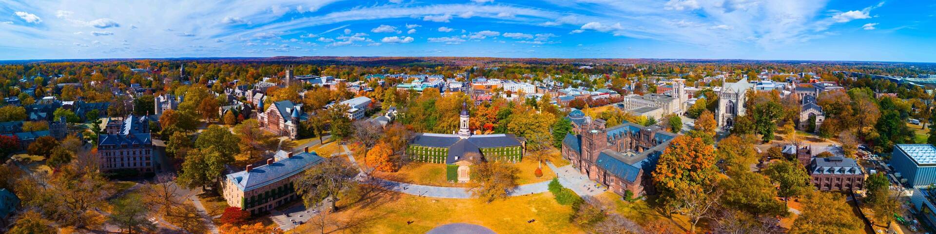 An aerial view of a university campus with historic buildings, a large green lawn, and autumn-colored trees.