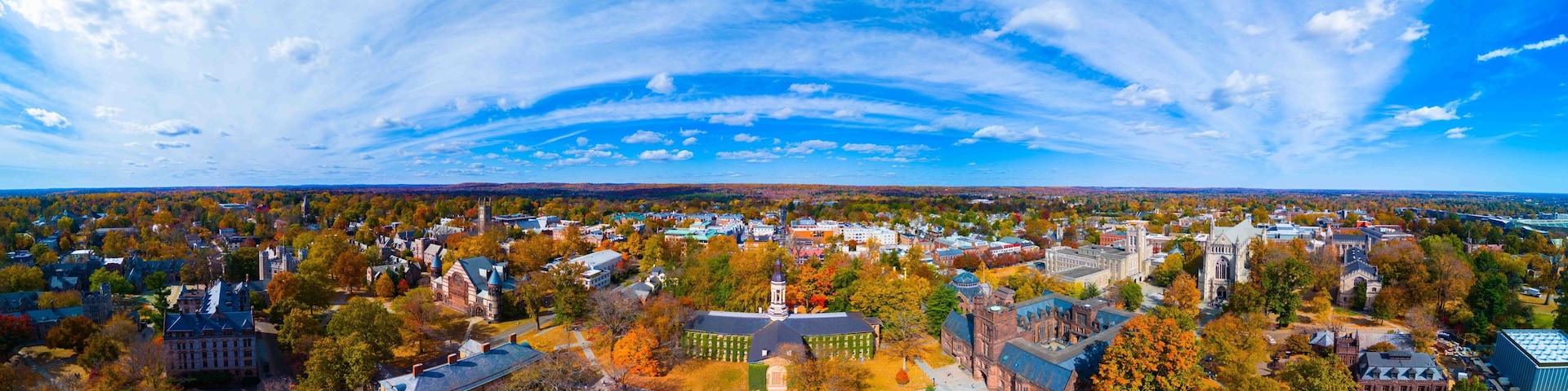 Vista aérea de um campus universitário com edifícios históricos, um gramado verde amplo e árvores em tons de outono.