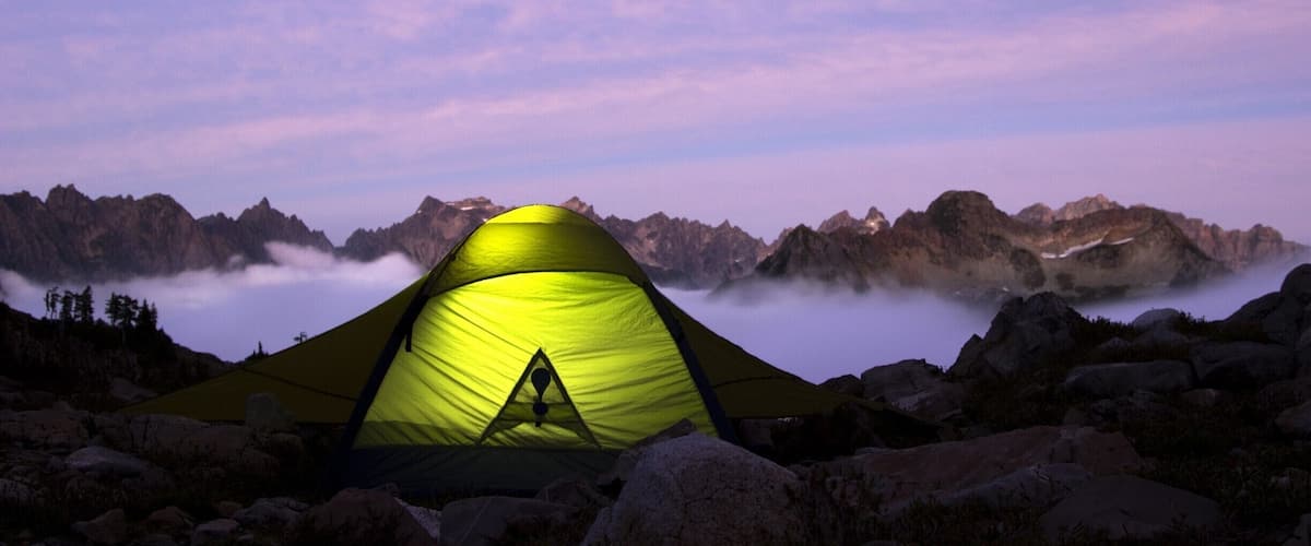 A yellow tent in a rocky mountainous area with a view of mist-covered peaks and a clear sky.