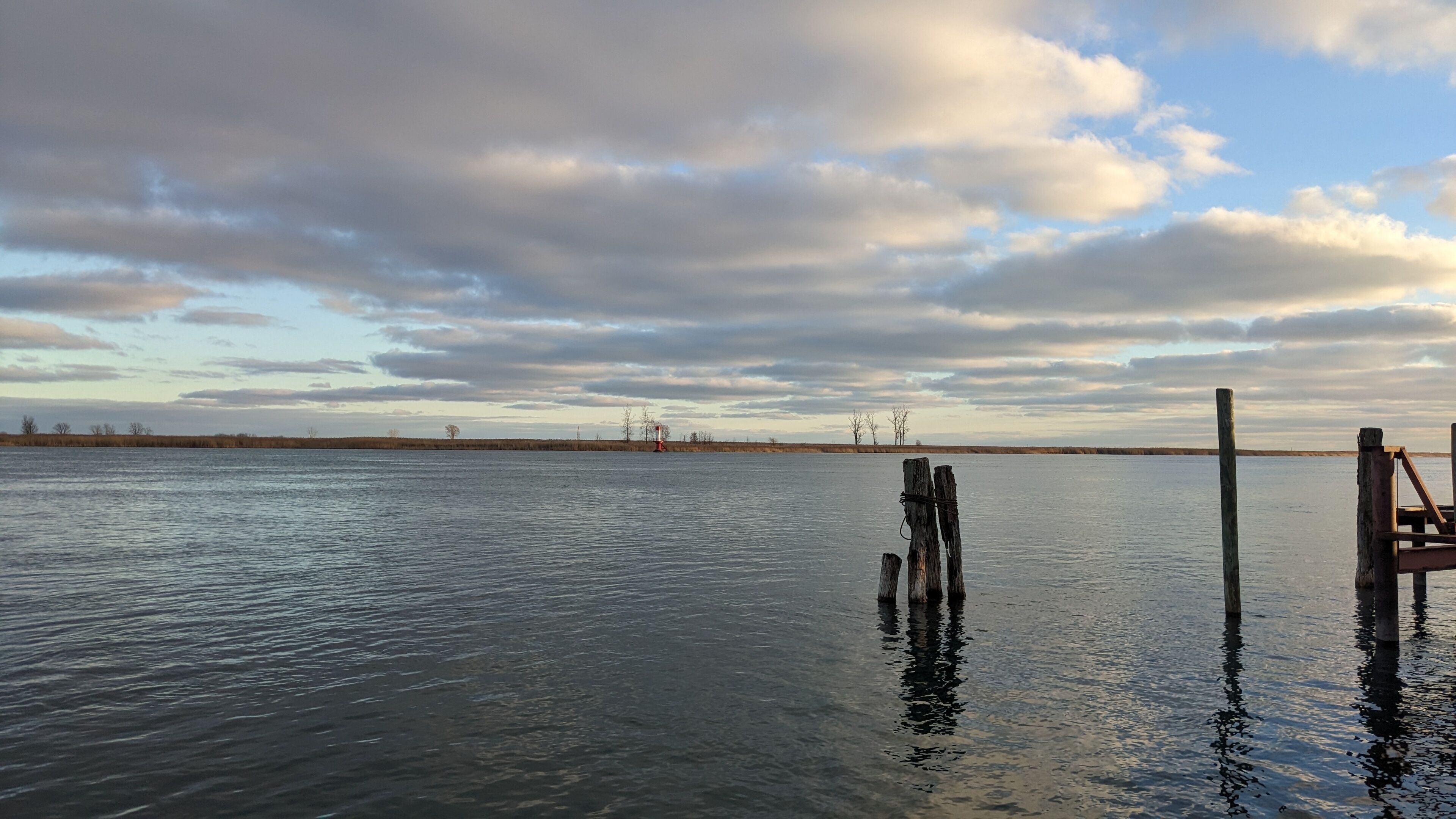 Un lago tranquilo con un muelle de madera que se adentra en el agua, rodeado de un paisaje llano y un cielo nublado.