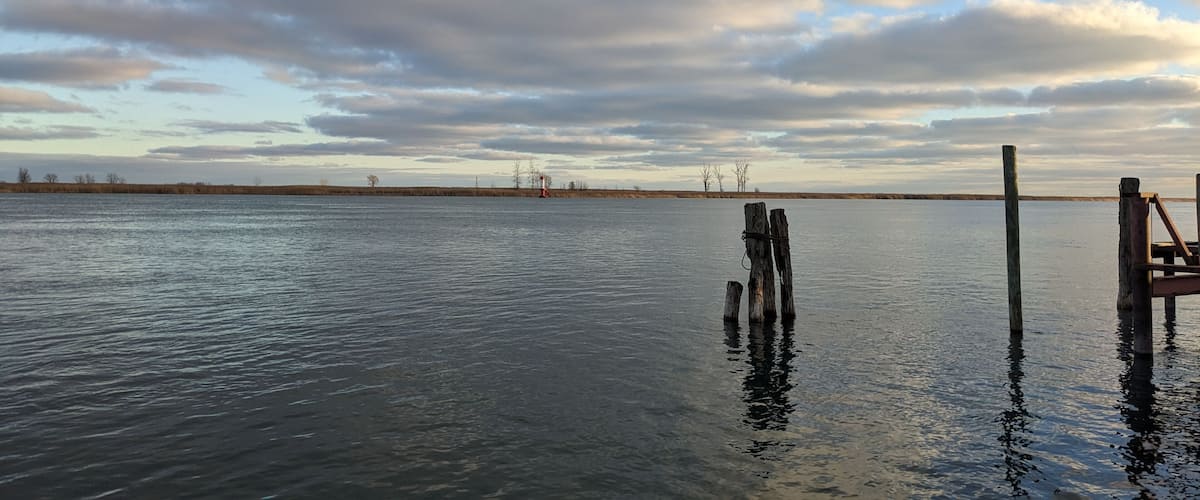 Un lac paisible, avec une jetée en bois qui s’avance sur l’eau, entouré d’un paysage plat et sous un ciel nuageux.