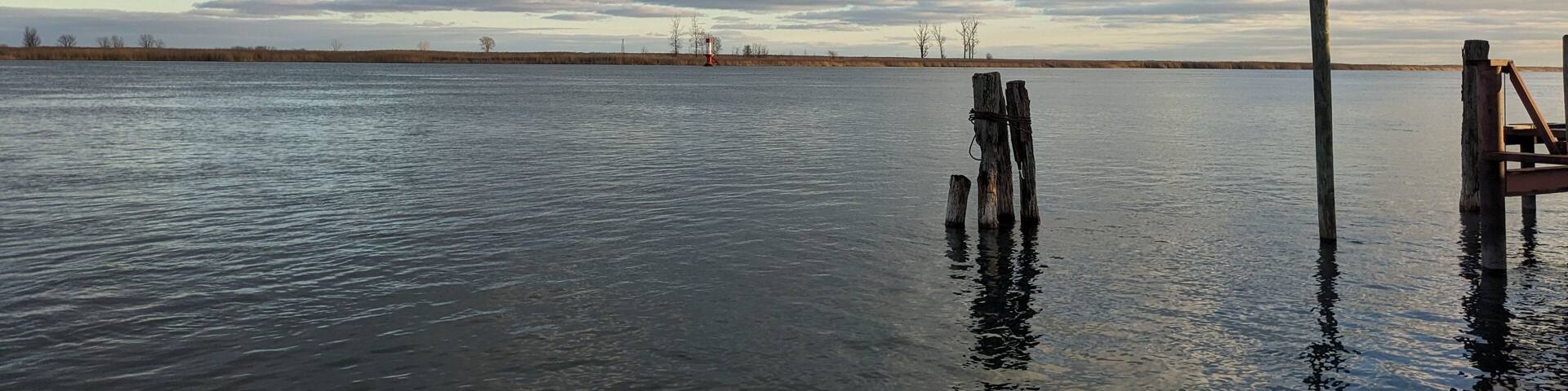 A calm lake with a wooden dock extending into the water, surrounded by a flat landscape and a cloudy sky.