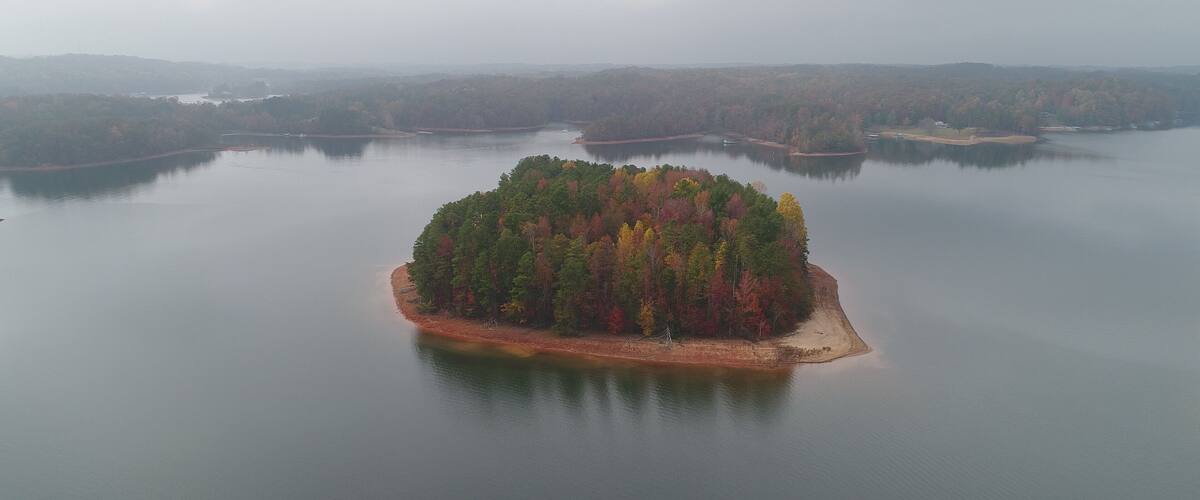An aerial view of a small island surrounded by calm water, with dense forested areas and distant hills.