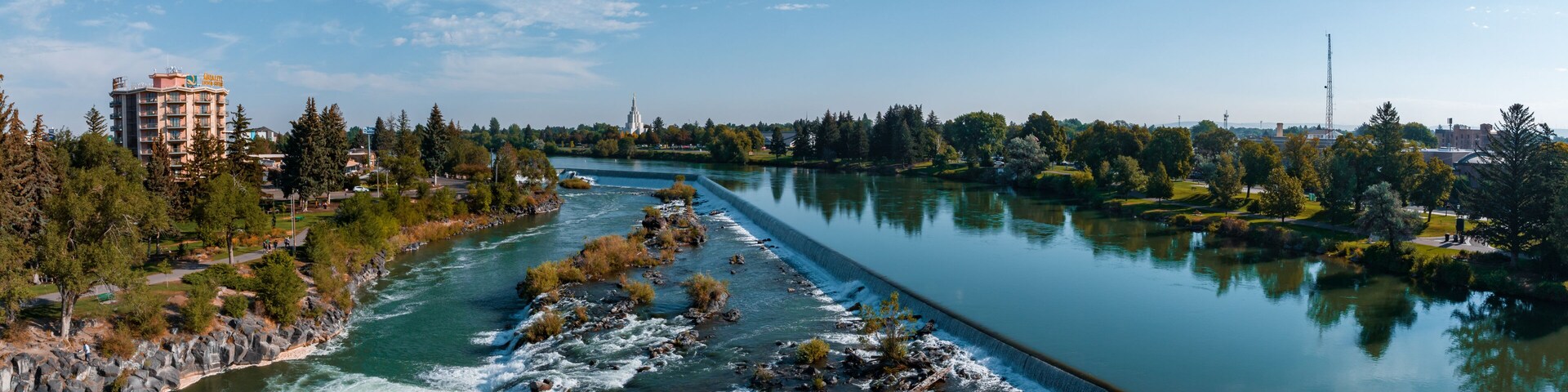 A river with a dam, a cityscape with buildings, and a clear blue sky.