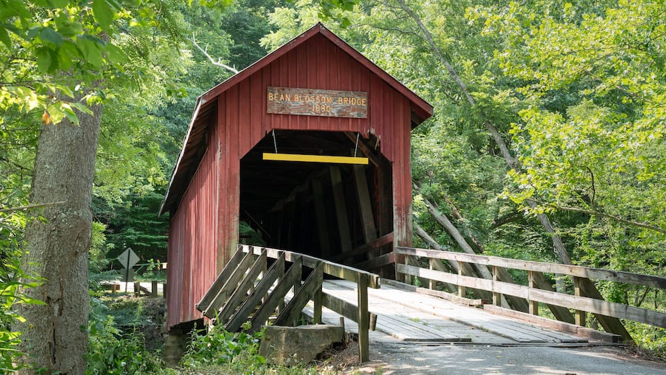 Un puente cubierto de color rojo con un cartel que dice "Bean Blossom Bridge 1880" en una zona boscosa.