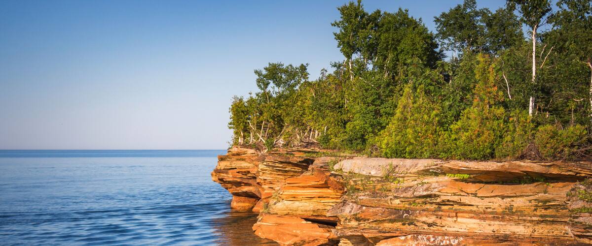 A rocky shoreline with clear blue water, dense green trees, and a clear sky.