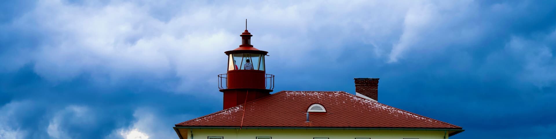 A white building with a red roof and a lighthouse tower, surrounded by tall grass and under a cloudy sky.