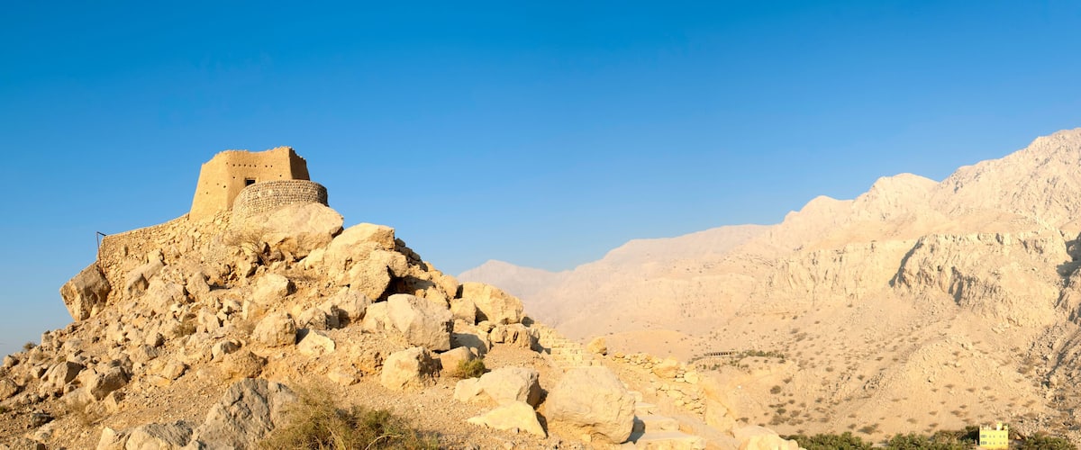 A rocky mountain with a small fortification on top and a large, barren hill in the background.