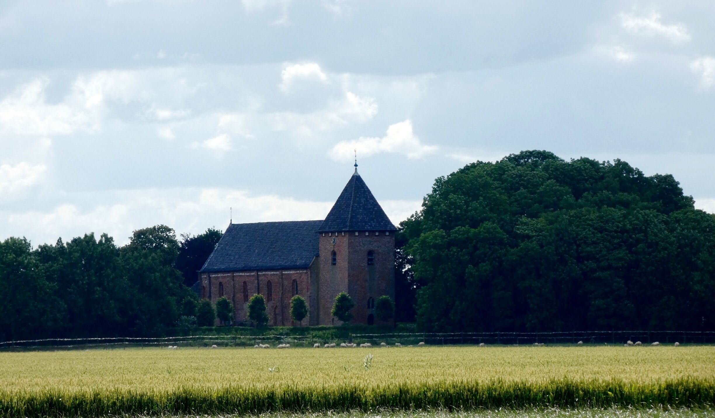 A historic building with a pointed roof and a clock tower, surrounded by lush greenery and a field.