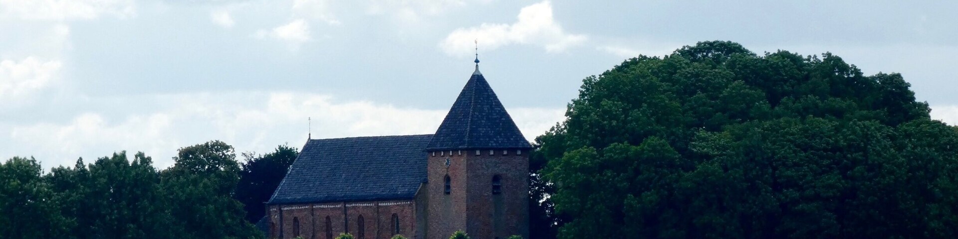 A historic building with a pointed roof and a clock tower, surrounded by lush greenery and a field.