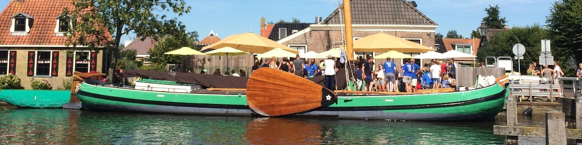 A traditional sailing boat docked at a waterfront with people on board and a building with a tiled roof in the background.