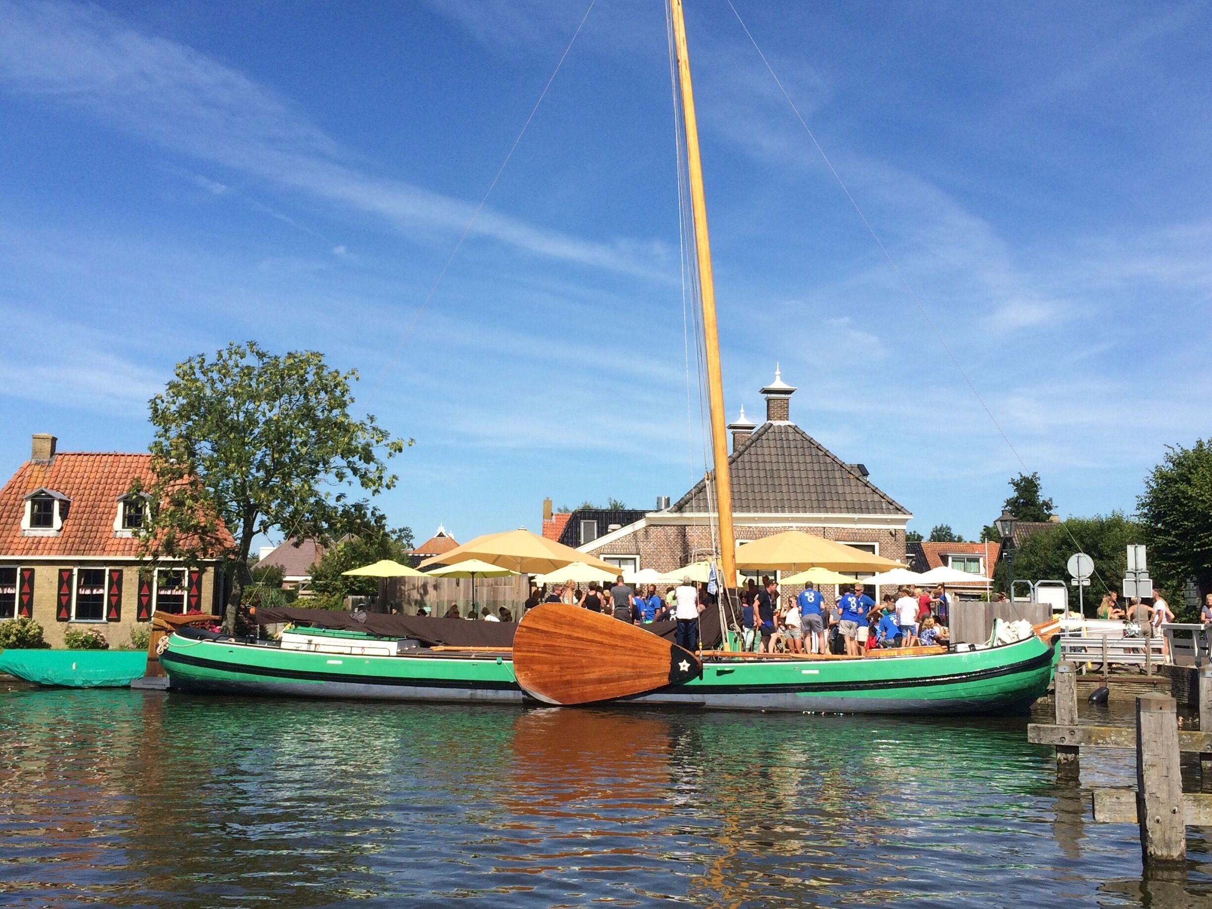 A traditional sailing boat docked at a waterfront with people on board and a building with a tiled roof in the background.