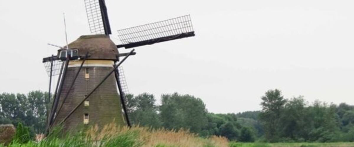 A windmill by a calm river with tall grass and a boat in the distance.