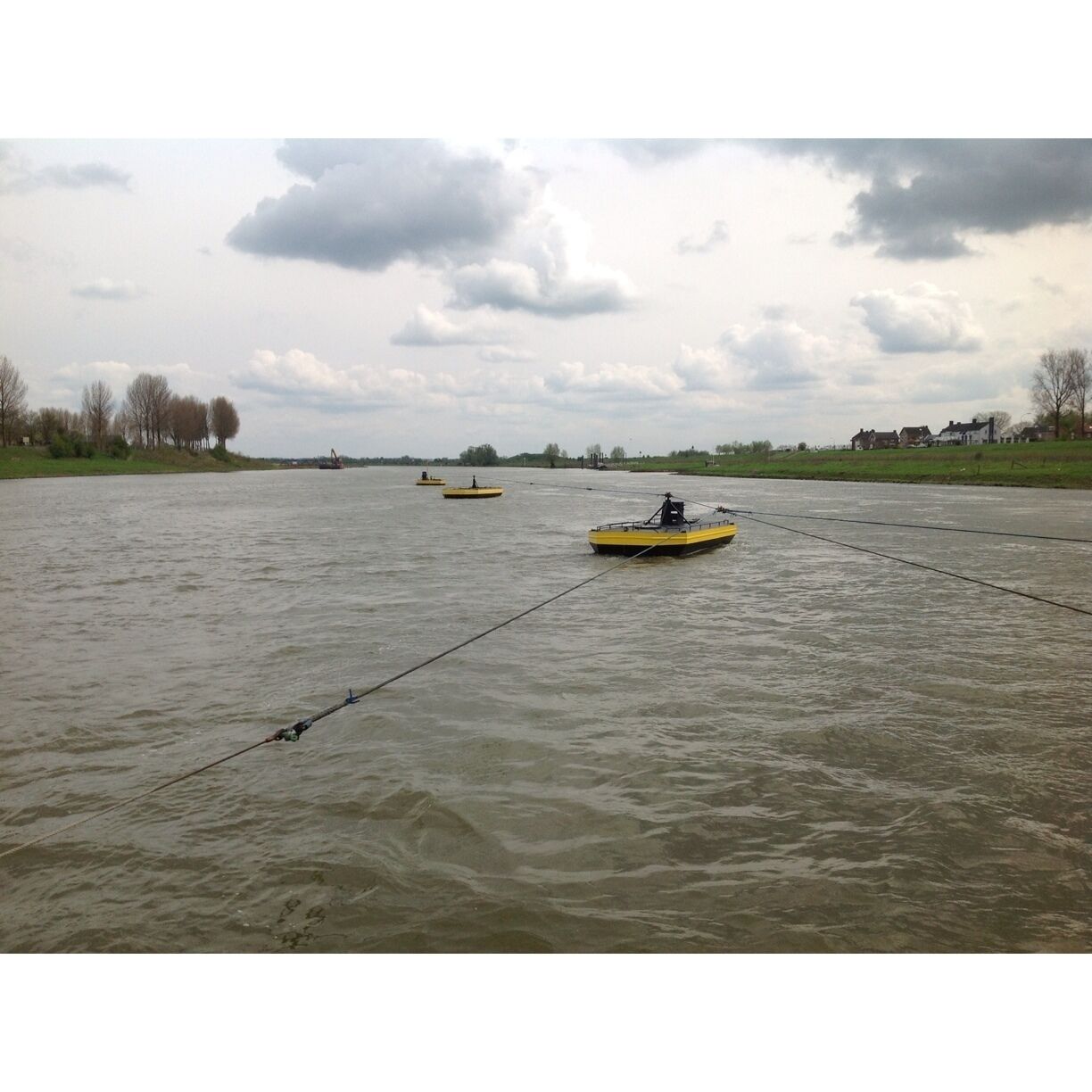 A river with three yellow boats, a cloudy sky, and houses in the distance.