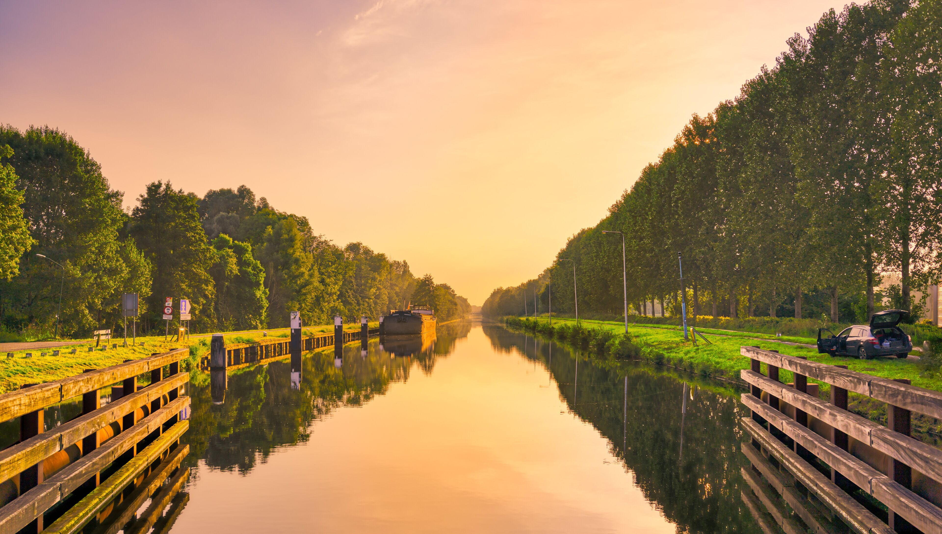 A calm river with a boat, surrounded by lush greenery and a wooden bridge.