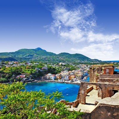 A coastal town with a historic fort, clear blue sea, and green hills in the background.
