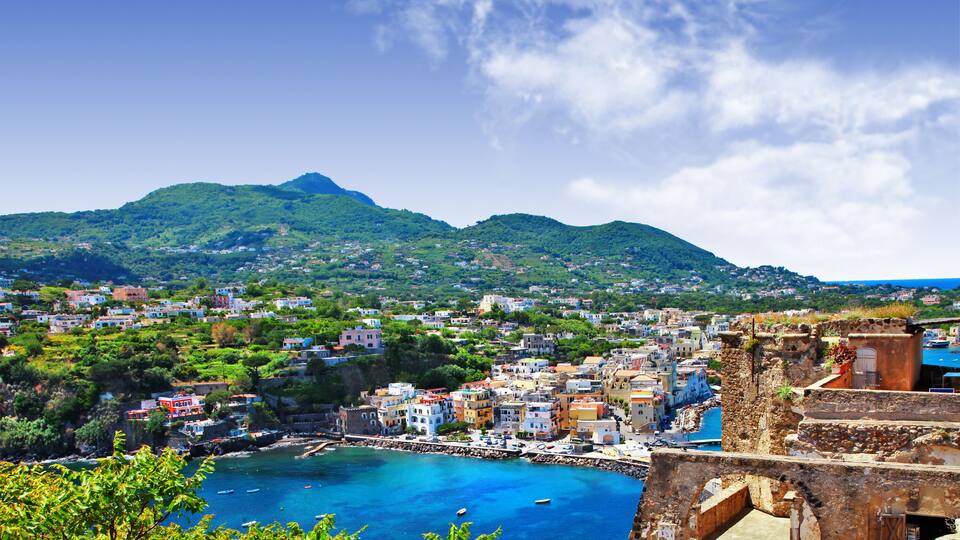A coastal town with a historic fort, clear blue sea, and green hills in the background.