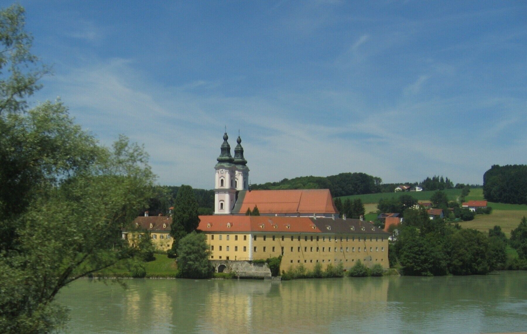 A historic building with a tower and red roof, surrounded by greenery and a calm lake.