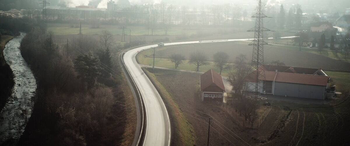 A winding road curving through a rural landscape with a river on one side, surrounded by fields and sparse trees.