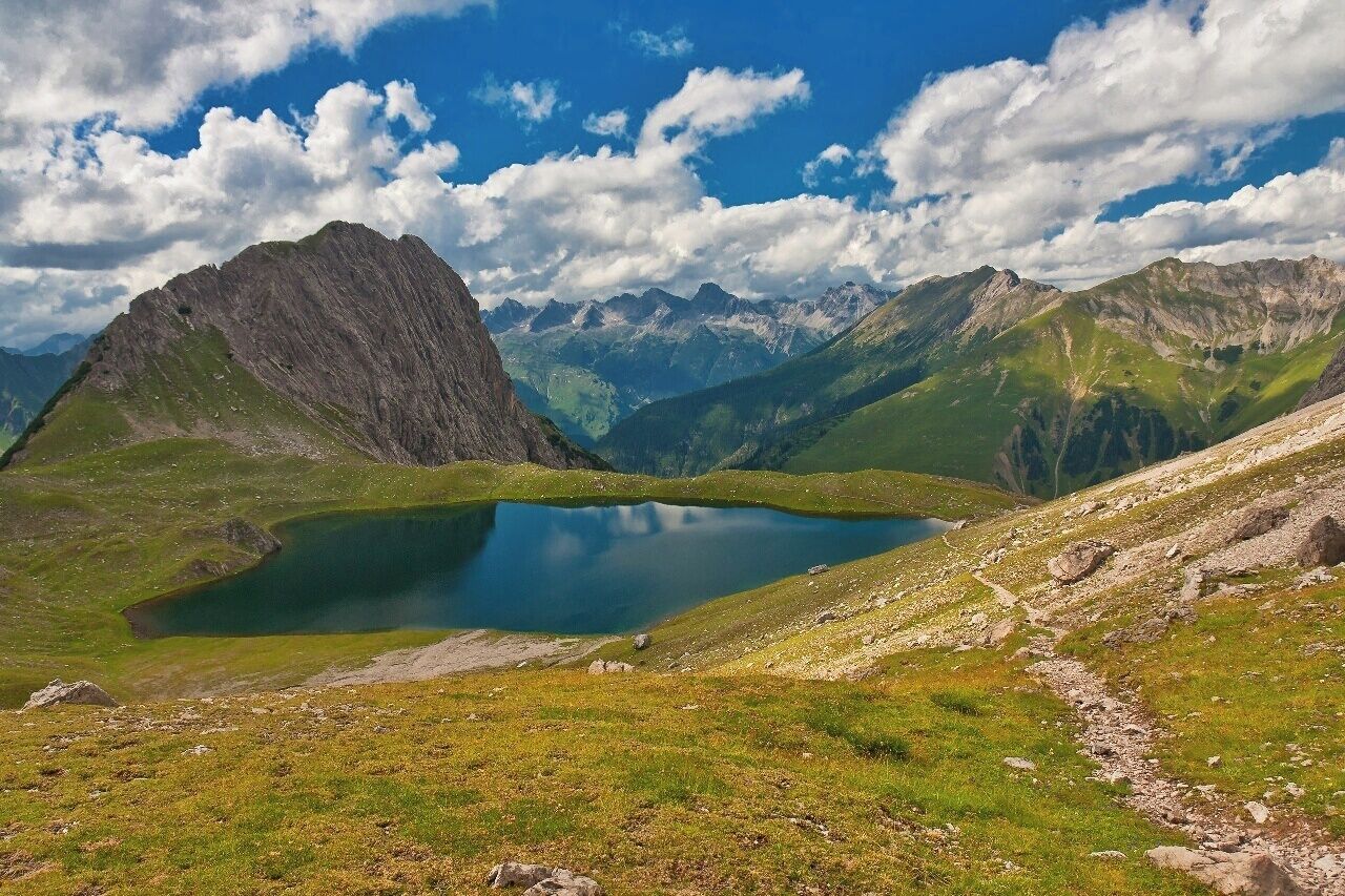 A mountain landscape with a clear lake, rocky terrain, and distant peaks under a partly cloudy sky.