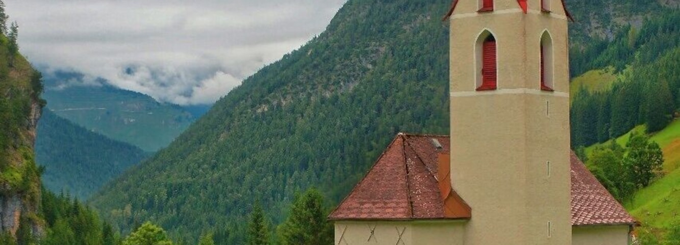 Una iglesia con un campanario y una aguja roja, rodeada de colinas verdes y un cielo nublado.