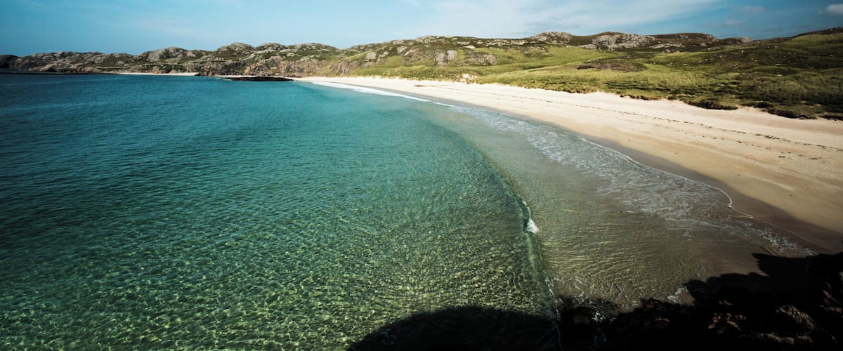 A coastal landscape with clear turquoise waters, a sandy beach, and rocky hills in the background.