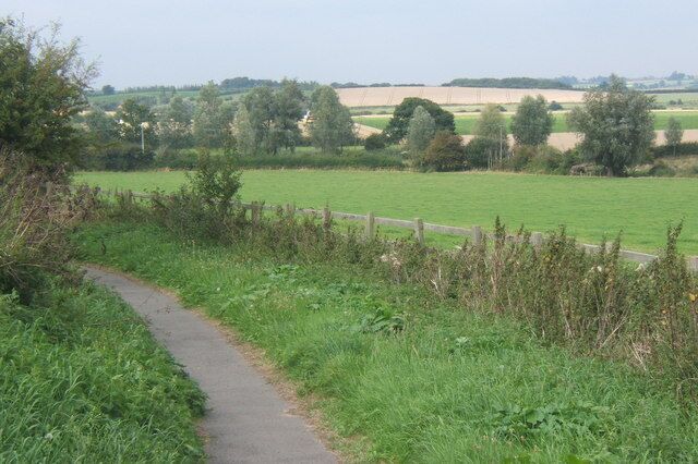 A rural path with a wooden fence, green fields, and trees in the distance.