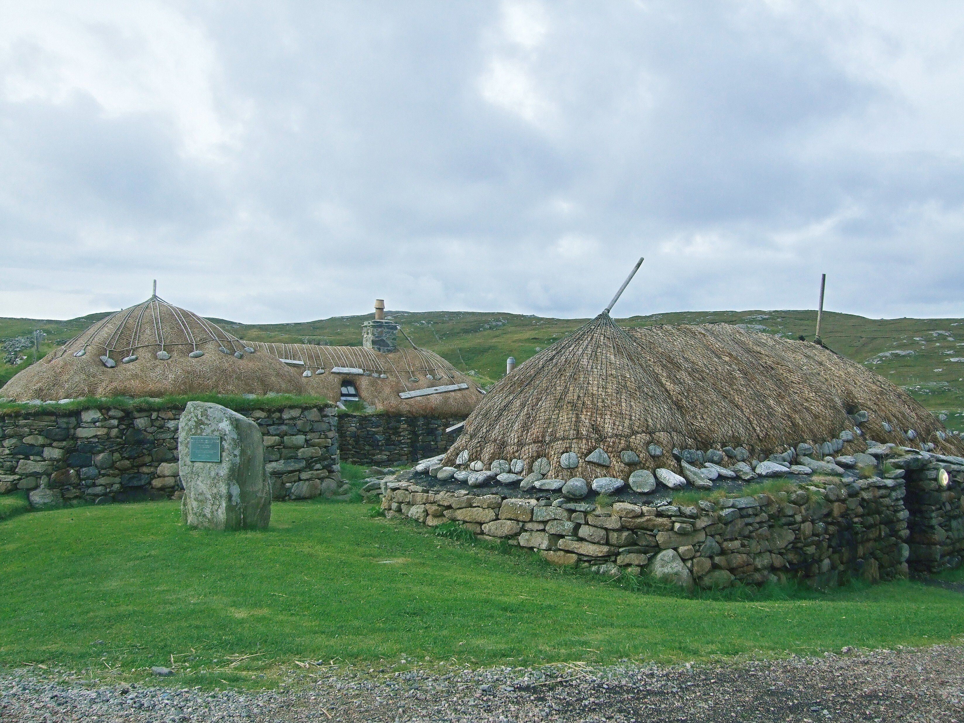 Traditional thatched-roof houses with stone walls and a stone cross in a grassy field.