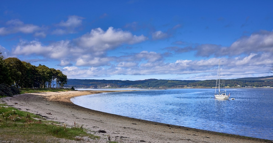 Un lago tranquilo con un velero, una playa arenosa y un promontorio boscoso al fondo.
