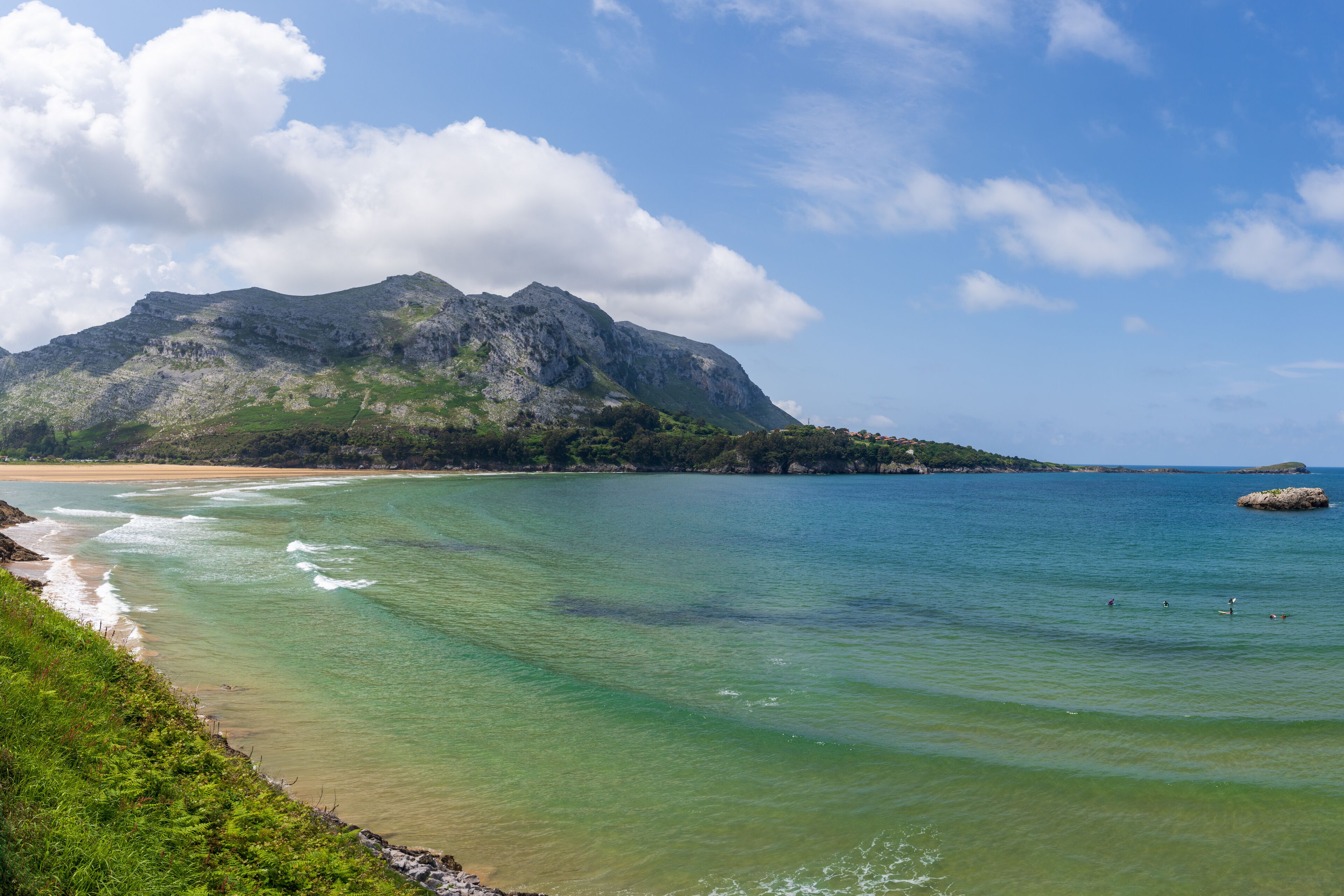 A coastal landscape with clear turquoise waters, a sandy beach, and a prominent mountain in the background.