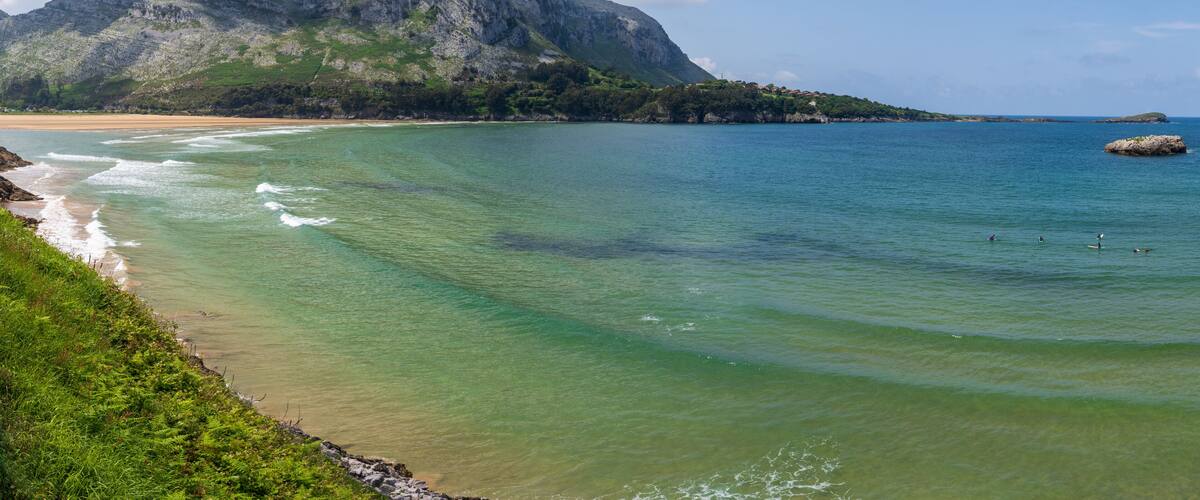 A coastal landscape with clear turquoise waters, a sandy beach, and a prominent mountain in the background.