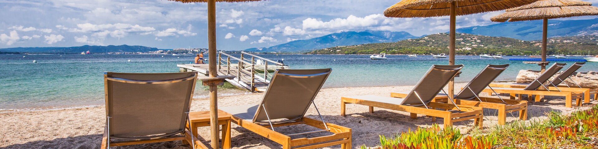 A beach with lounge chairs and thatched umbrellas, a pier, and a boat in the background.